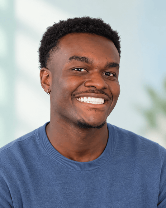 Front-facing portrait of a young man smiling naturally against a clean background with subtle mint and blue tones